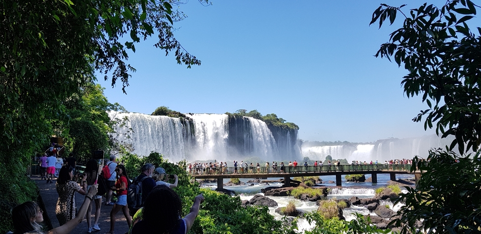 Tourists on a walkway with Iguazu Falls in the background on a clear day.