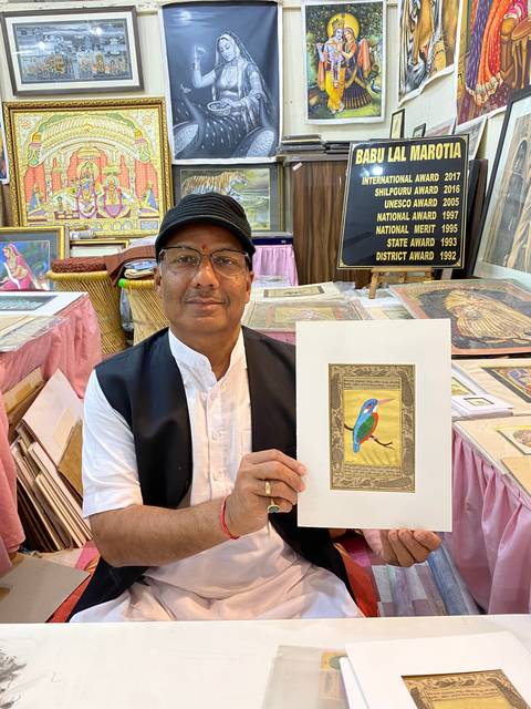       Man displaying artwork with various paintings in the background.
  