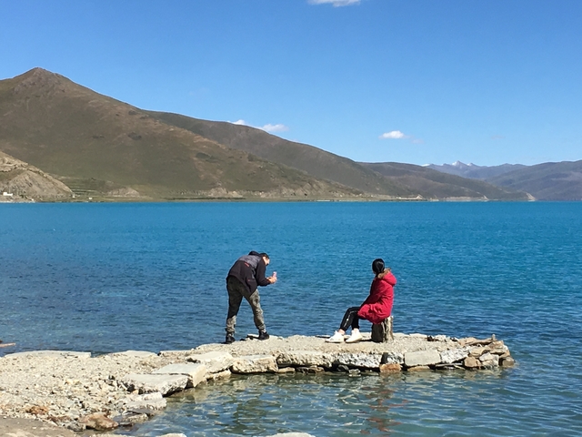 A person taking a photo of someone sitting by a clear blue lake with mountains.