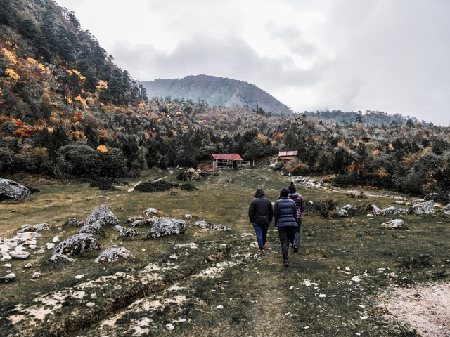       Two people walking towards traditional cabins surrounded by colorful autumn forest.
  