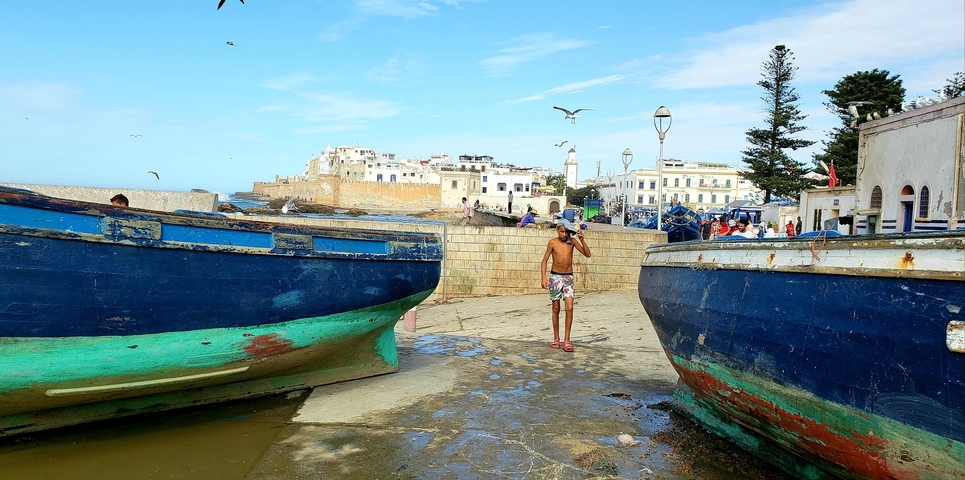       A person standing between two blue boats near a coastal town.
  