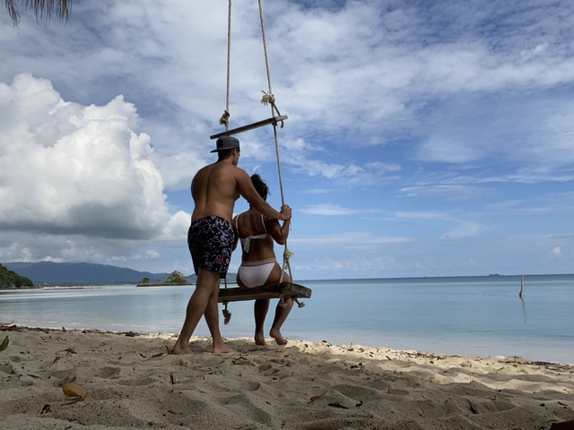       Couple on a swing by a tropical beach looking at the horizon.
  