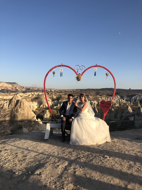       Couple in wedding attire sitting in front of a heart-shaped frame overlooking Cappadocia.
  