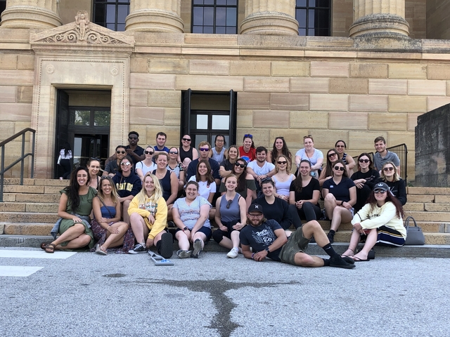 Large group sitting on steps in front of a sandstone building.