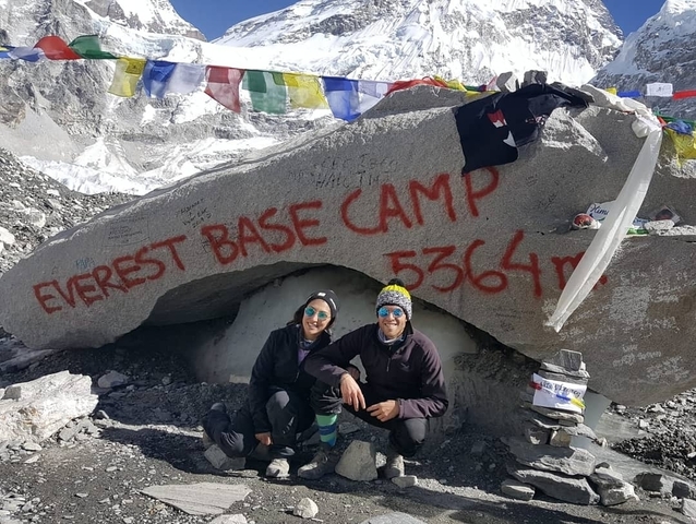       Two people posing at Everest Base Camp with flags.
  