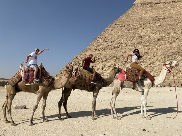       Three people riding camels near a pyramid.
  