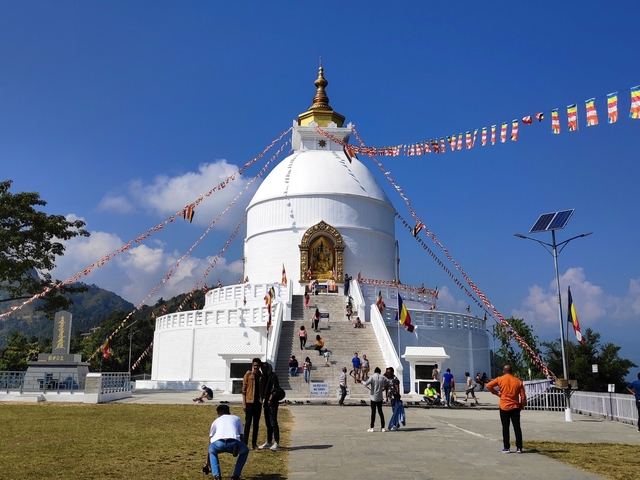       People climbing steps of a large white stupa with flags and blue sky.
  