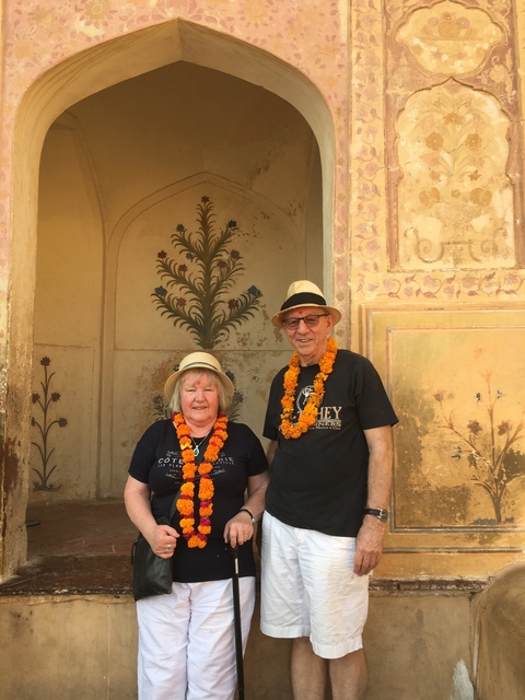 Two older adults with marigold garlands in front of a decorated wall.