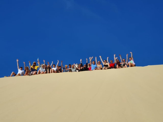       Group of people seated on a sand dune with hands raised.
  