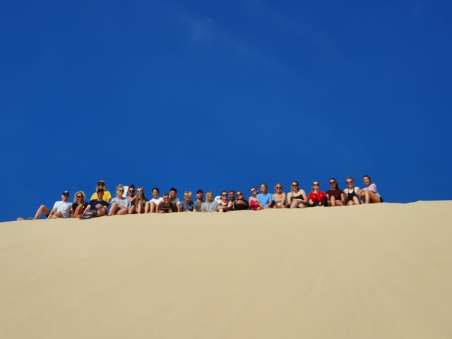      Group of people sitting on a large sand dune with a clear blue sky.
  