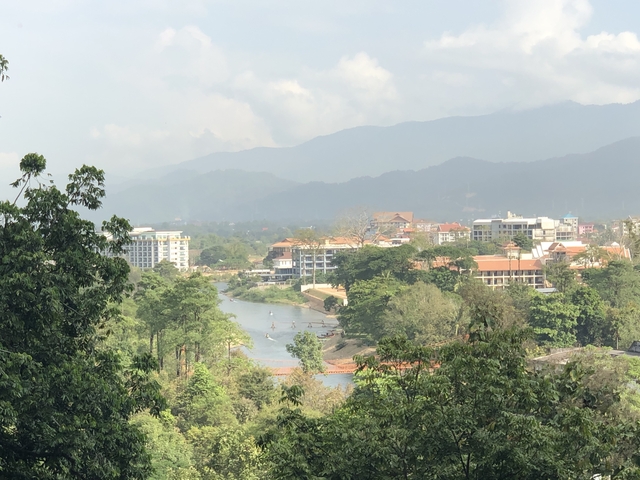       Village beside a river with mountains in the background under a cloudy sky.
  