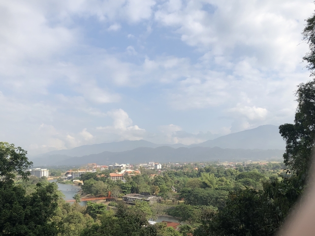       Wide view of a town by a river with mountain backdrop and cloudy sky.
  