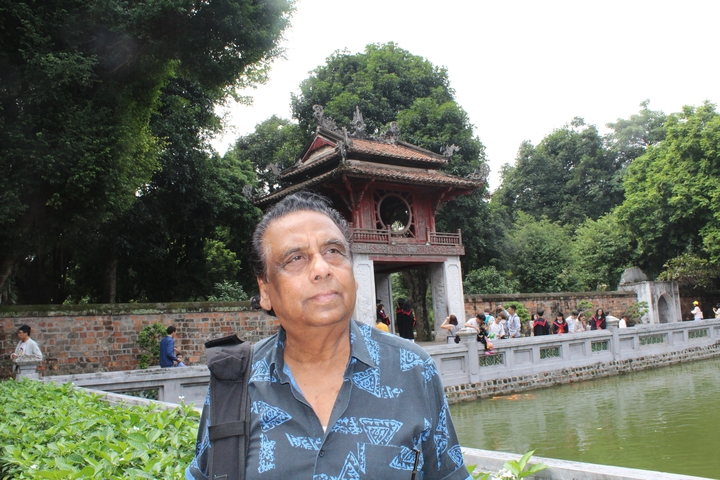 Person standing in front of a traditional structure with water and trees in the background.