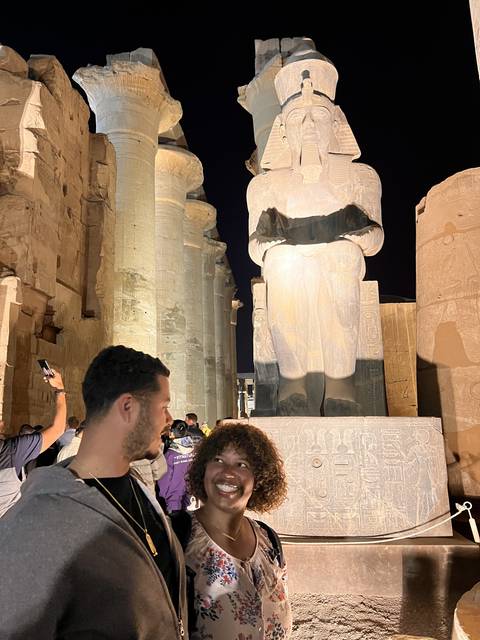      Two people smiling in front of ancient Egyptian statues at night.
  