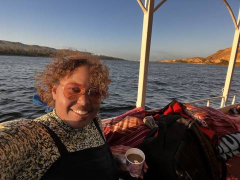      Person smiling on a boat with water and landscape.
  