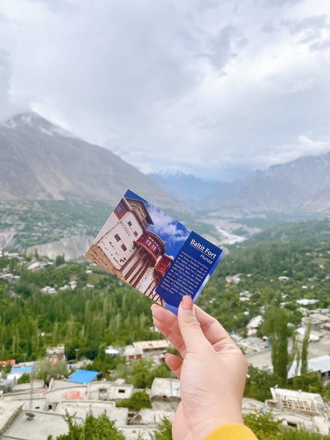       Hand holding a postcard of Baltit Fort with mountainous background.
  