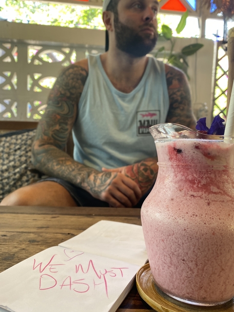A pink smoothie in a glass with a person having tattoos in the background.