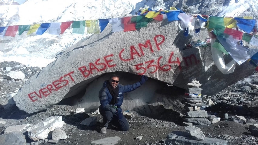       Person posing at Everest Base Camp with a marked rock and colorful flags.
  