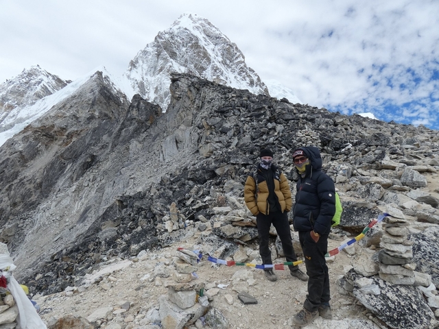 Two people in winter clothing standing on a rocky path with snowy mountains behind.