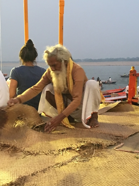       An elderly man sitting on a riverside, arranging brown fibers with people and boats in the background.
  