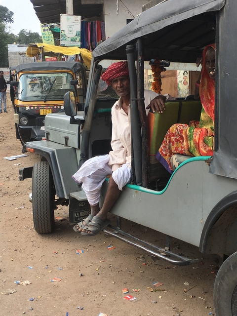 An elderly man sitting in a rustic vehicle with colorful fabric, parked in a dusty area.