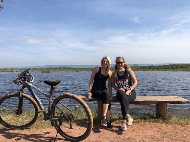       Two women sitting on a bench with a bicycle by a lake.
  