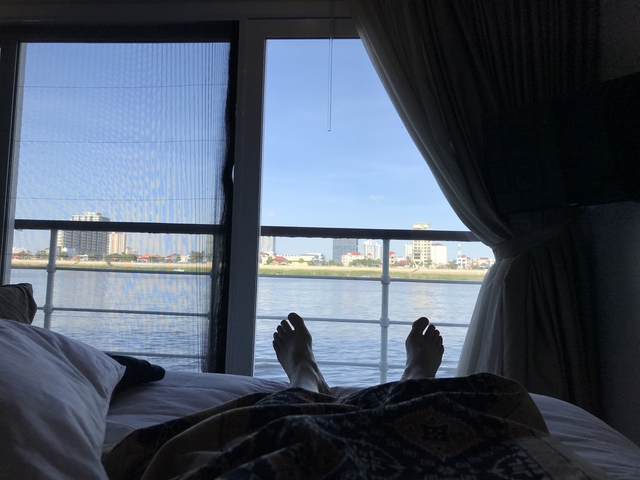       View from a bed through a large window of a city skyline and river.
  