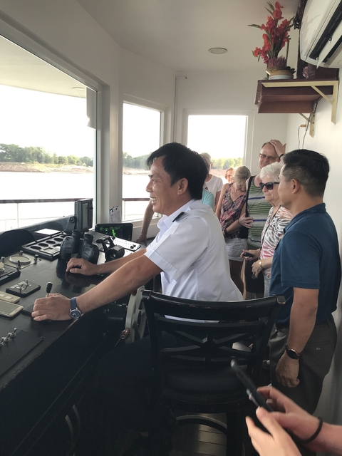 People interacting with a ship's captain in a wheelhouse.