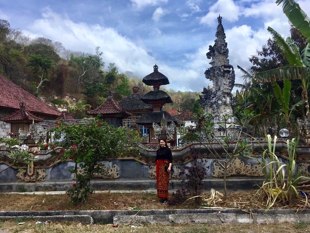       A person standing in front of traditional Indonesian architecture and lush greenery.
  