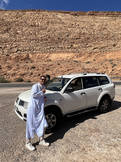       Man in traditional clothing standing by a parked SUV in a desert.
  
