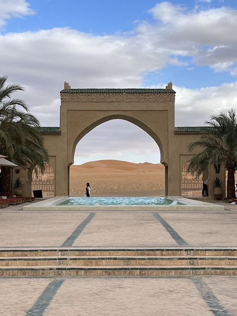       View of a pool with decorative arch leading to desert sand dunes.
  
