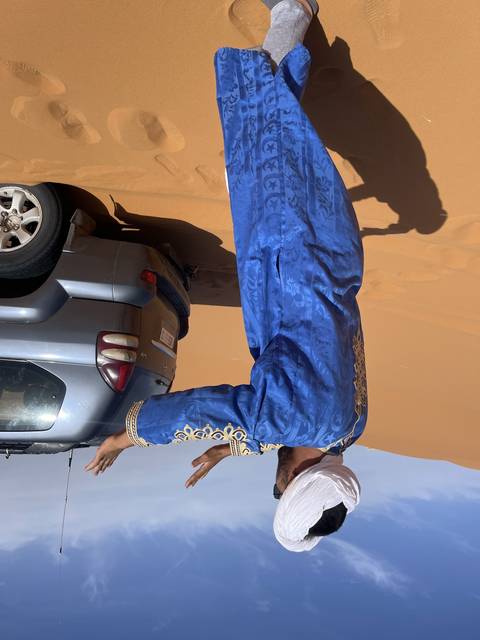 Person with traditional attire on sand dune near a vehicle.