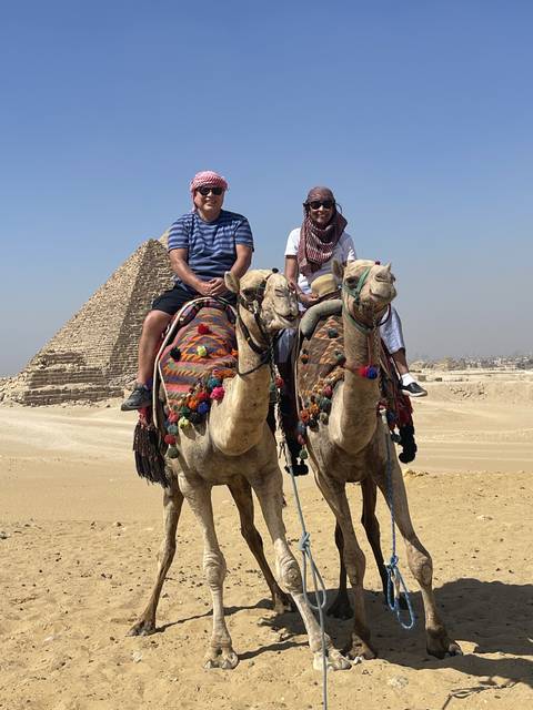       Two people riding camels with pyramids visible.
  
