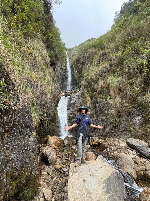 Person posing at a waterfall in a natural setting.