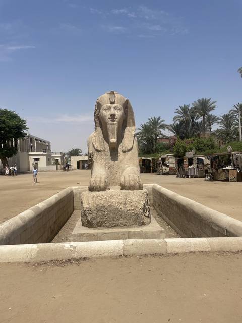       Stone statue of an Egyptian sphinx with palm trees in the background.
  
