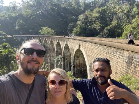 Three people posing near a bridge surrounded by trees.