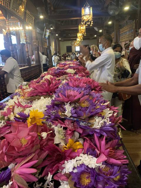 A vibrant collection of colorful flowers with people in the background, likely in a market or temple setting.