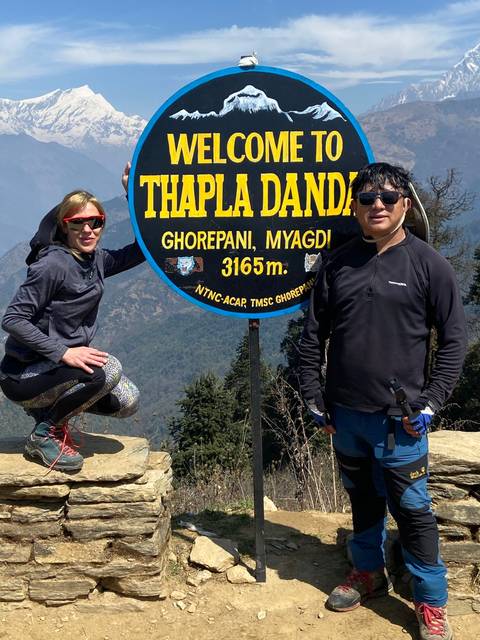 People posing with a sign saying 'Welcome to Thapla Danda' with snowy mountains in the background.