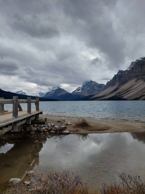 A lake with a small wooden dock, surrounded by mountains under a cloudy sky.