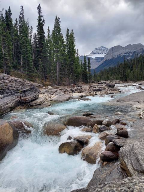       Flowing river with rocky banks and trees, surrounded by mountains.
  