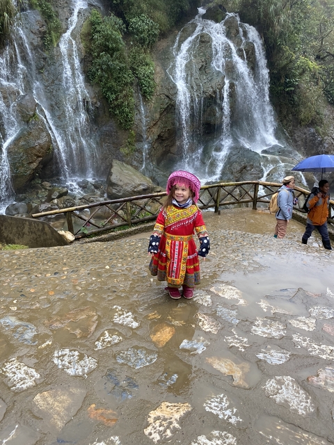       Child in traditional attire by a waterfall and tourists exploring.
  
