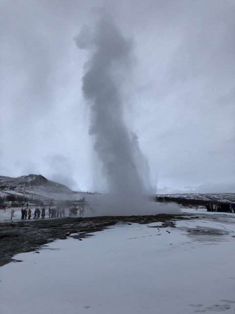 A geyser erupting with people watching in the background on a snowy landscape.