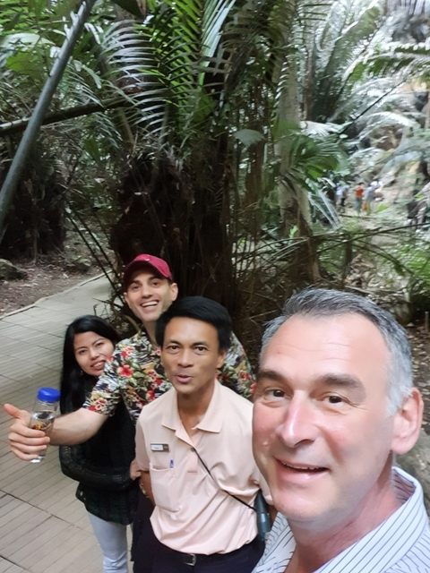 Group of people smiling, standing on a wooden path in a forest.