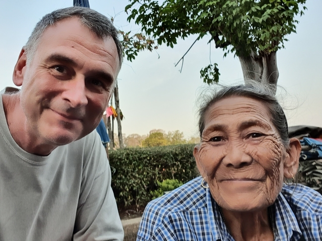 Two people smiling, one elderly, with trees in the background.