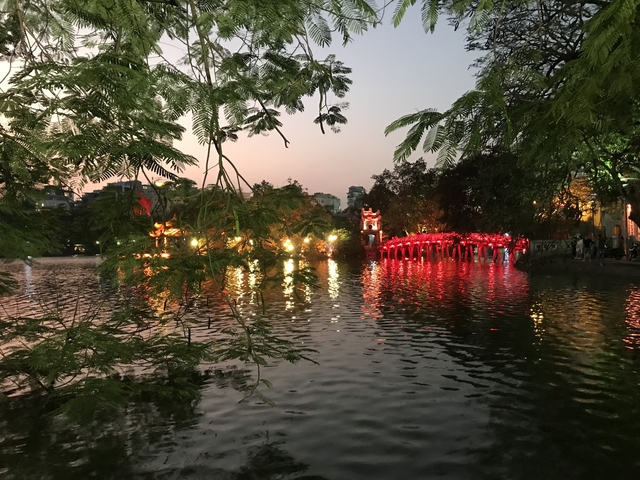       Lit-up red bridge over water, surrounded by trees at dusk.
  