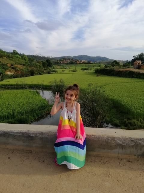 A child posing with a peace sign in front of lush green rice paddies and distant hills.