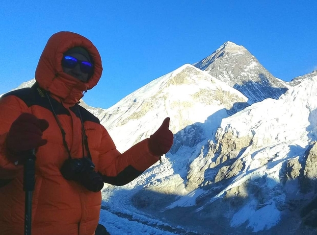 A person in winter gear with reflective sunglasses giving a thumbs up in a snowy mountain landscape.