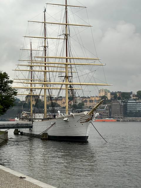 Sailing ship docked near a waterfront with city buildings.