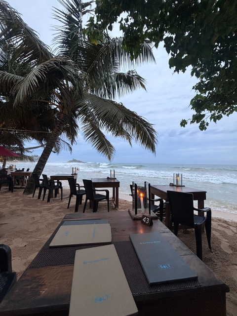       Beachside dining area with tables and chairs under palm trees.
  