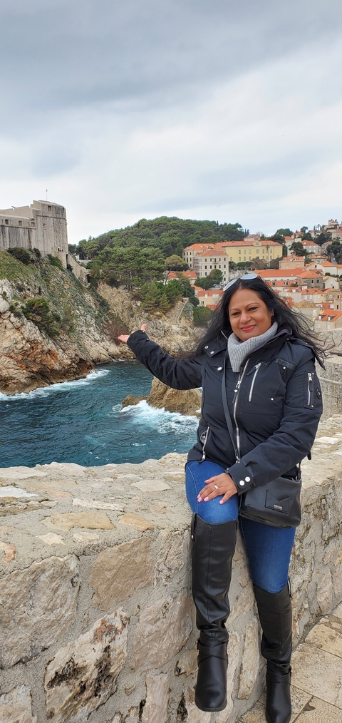Person posing by the sea with a view of rocky cliffs and historic buildings.